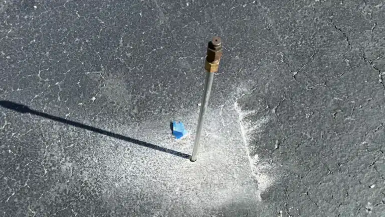Metal probe inserted into cracked asphalt with light debris around the hole, used for measuring ground conditions.