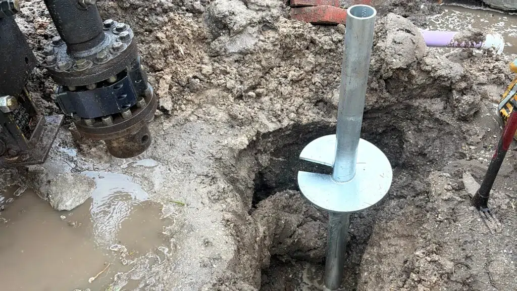 Construction site with a muddy excavation where a metal helical pier is installed vertically into the ground using hydraulic drilling equipment