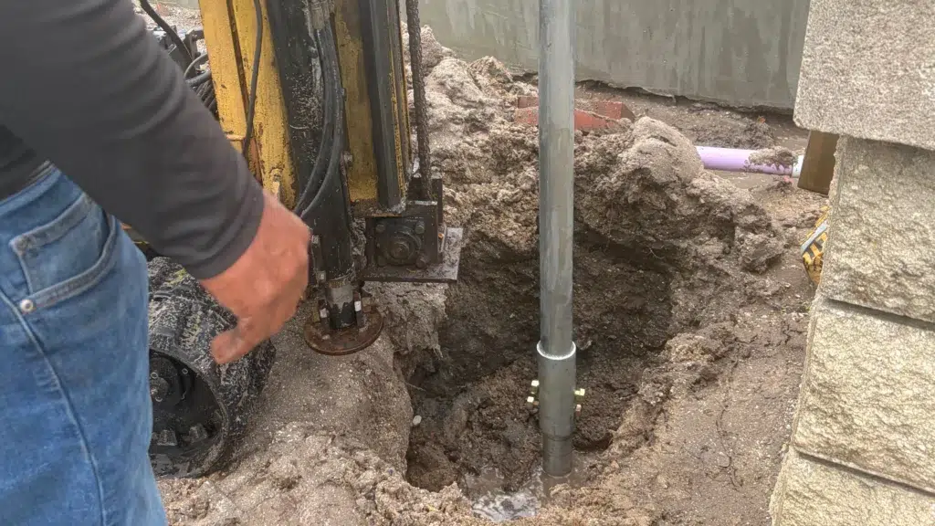 Worker operating hydraulic equipment next to a deep, narrow excavation where a steel helical pier shaft has been driven into wet soil