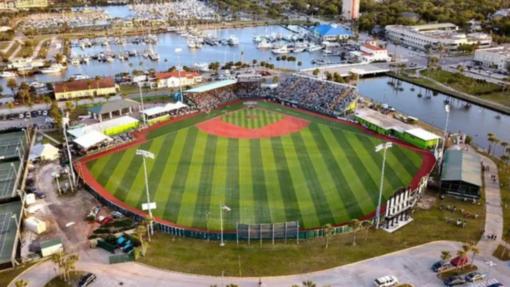 Aerial view of a baseball stadium with green striped outfield grass, surrounded by seating, marina water, boats, and nearby buildings