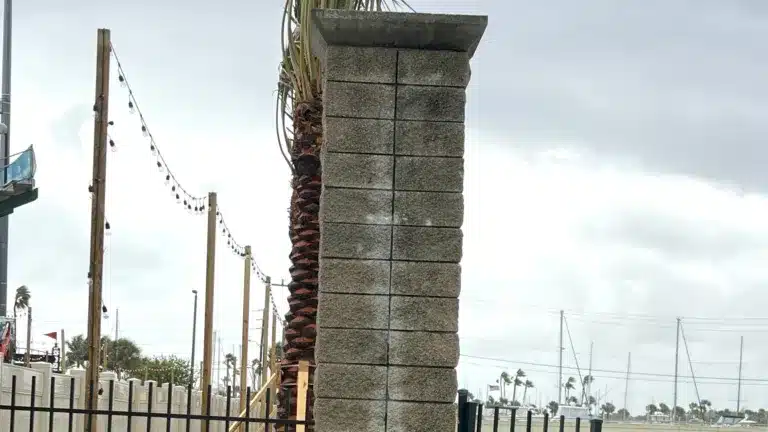 Tall rectangular stone column beside a waterfront walkway, with string lights, palm trees, marina masts, and an overcast sky in the background.