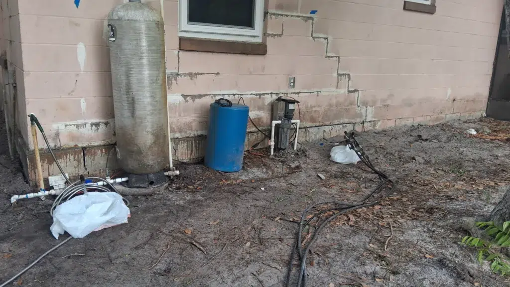 Outdoor well pump setup with pressure tanks, exposed PVC plumbing, and visible cracks on the home’s exterior wall