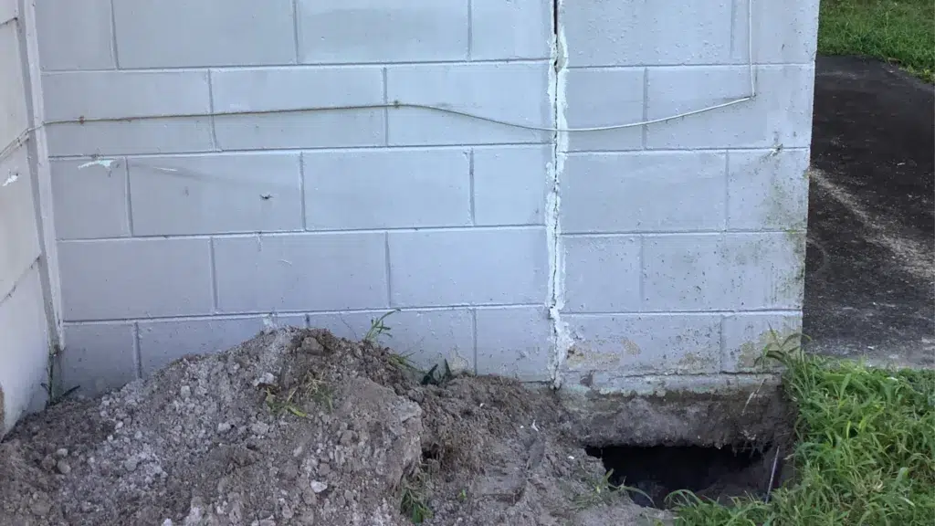Residential foundation wall with vertical cracking and excavation beneath the structure, exposing soil and footing area during foundation repair assessment.