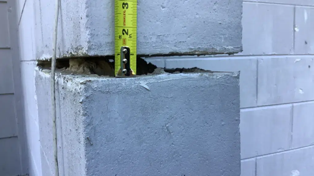 Measuring the width of a horizontal gap between displaced concrete block sections in a residential foundation wall using a tape measure to document structural separation.