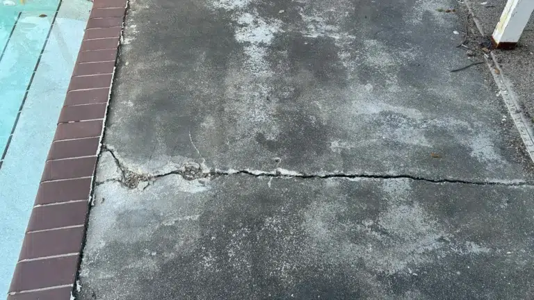 Residential concrete pool deck with a large horizontal crack and surface deterioration next to brick coping and swimming pool.