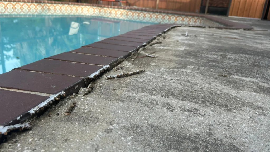 Damaged residential pool deck with lifted concrete slab, eroded base, and separation along brick coping near swimming pool.